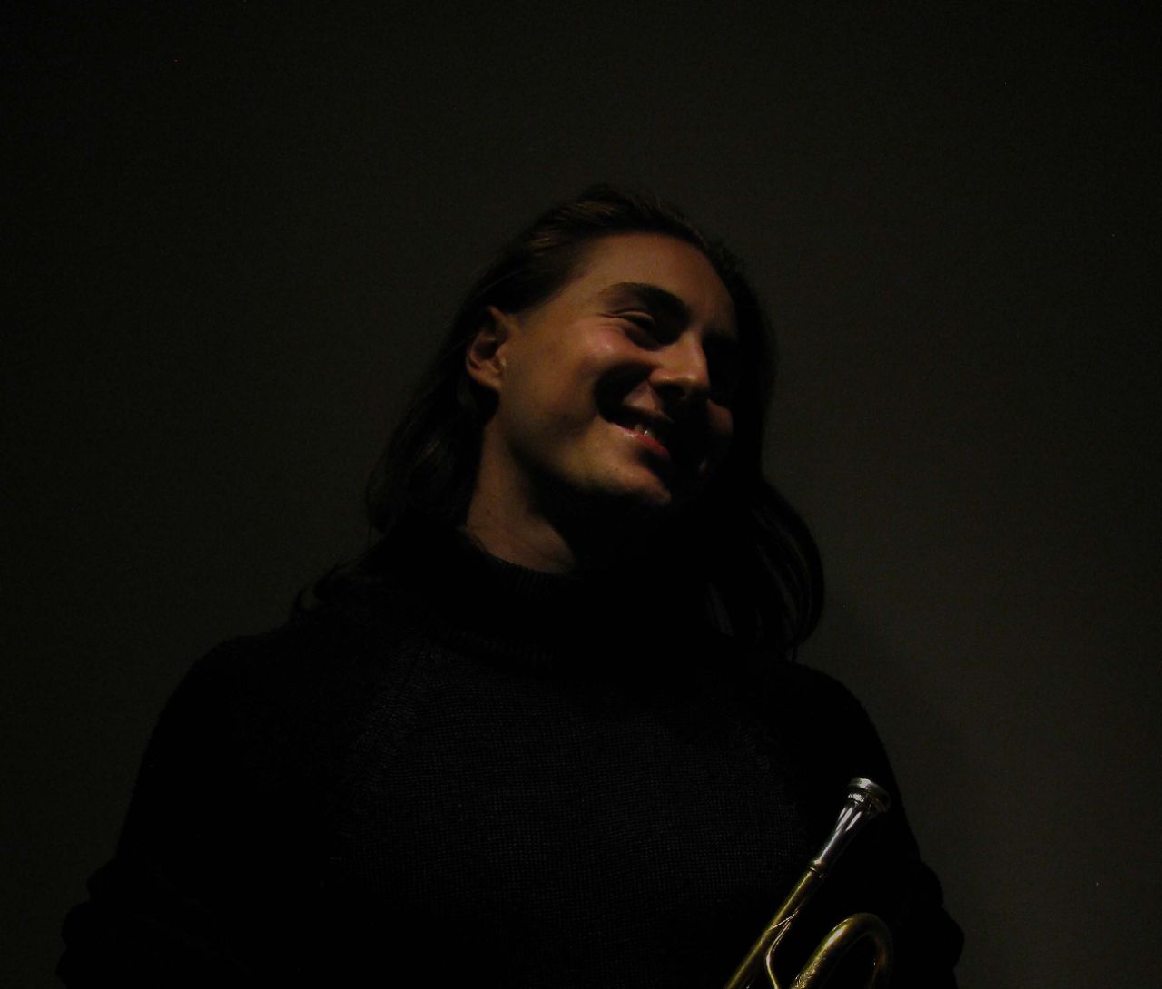 Valerian Alfaré mit seiner Trompete. Er lächelt und trägt lange, dunkle Haare und ein schwarzes Shirt. 